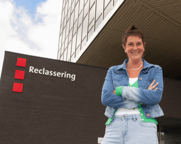 Vrouwelijke collega staat glimlachend voor een gebouw van Reclassering Nederland met het logo erop