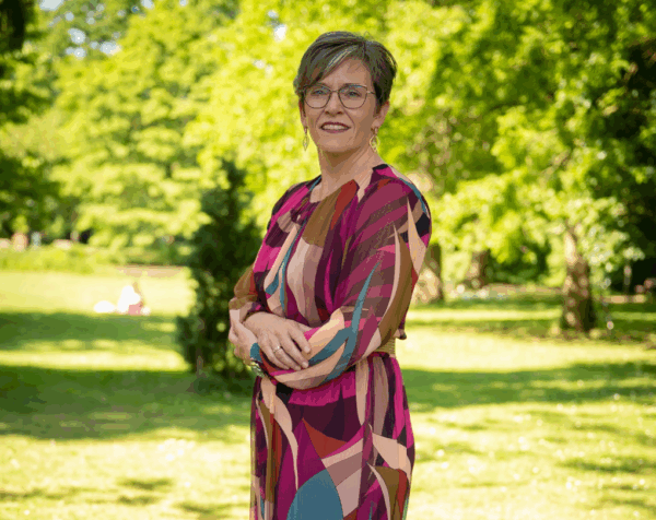 directielid Monique van Walt-Praag staand met handen over elkaar in het park, gekleed in een rode jurk met print.