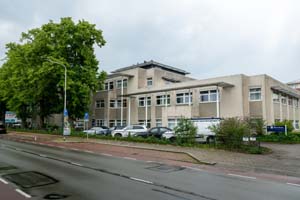 beige/creme gebouw langs de weg, met auto's schuin parallel geparkeerd en bomen links