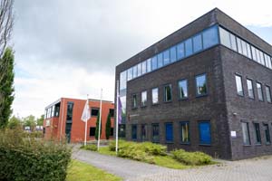 Donkergrijs gebouw met drie verdiepingen, drie grote vlaggen voor het voetenpad met struiken ernaast