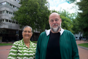 Twee reclasseringswerkers poseren voor een een gebouw. Links een vrouw met groen-wit blouse en rechts een man met baard en groen vest.